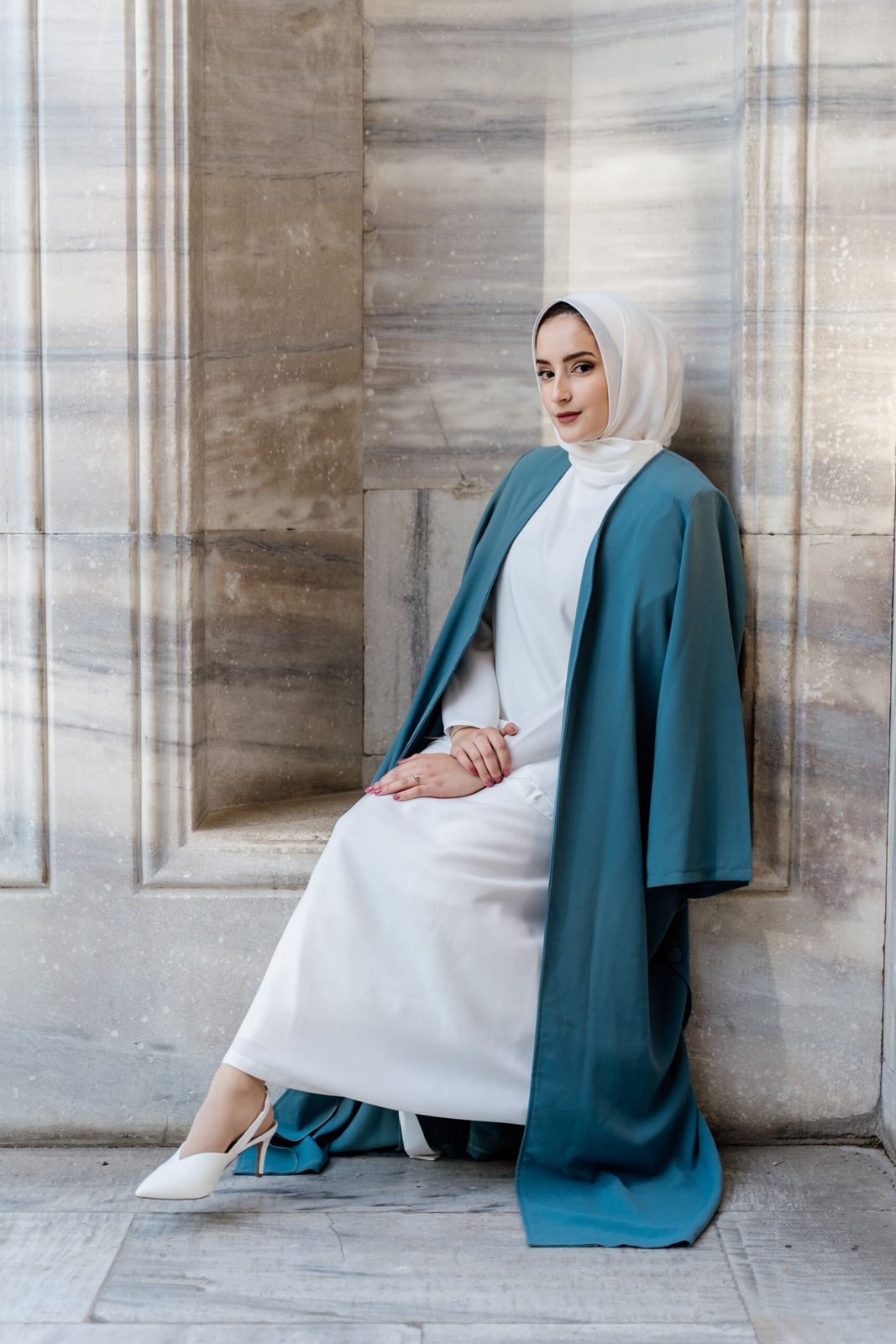 Woman in a white dress and teal coat seated beside marble columns