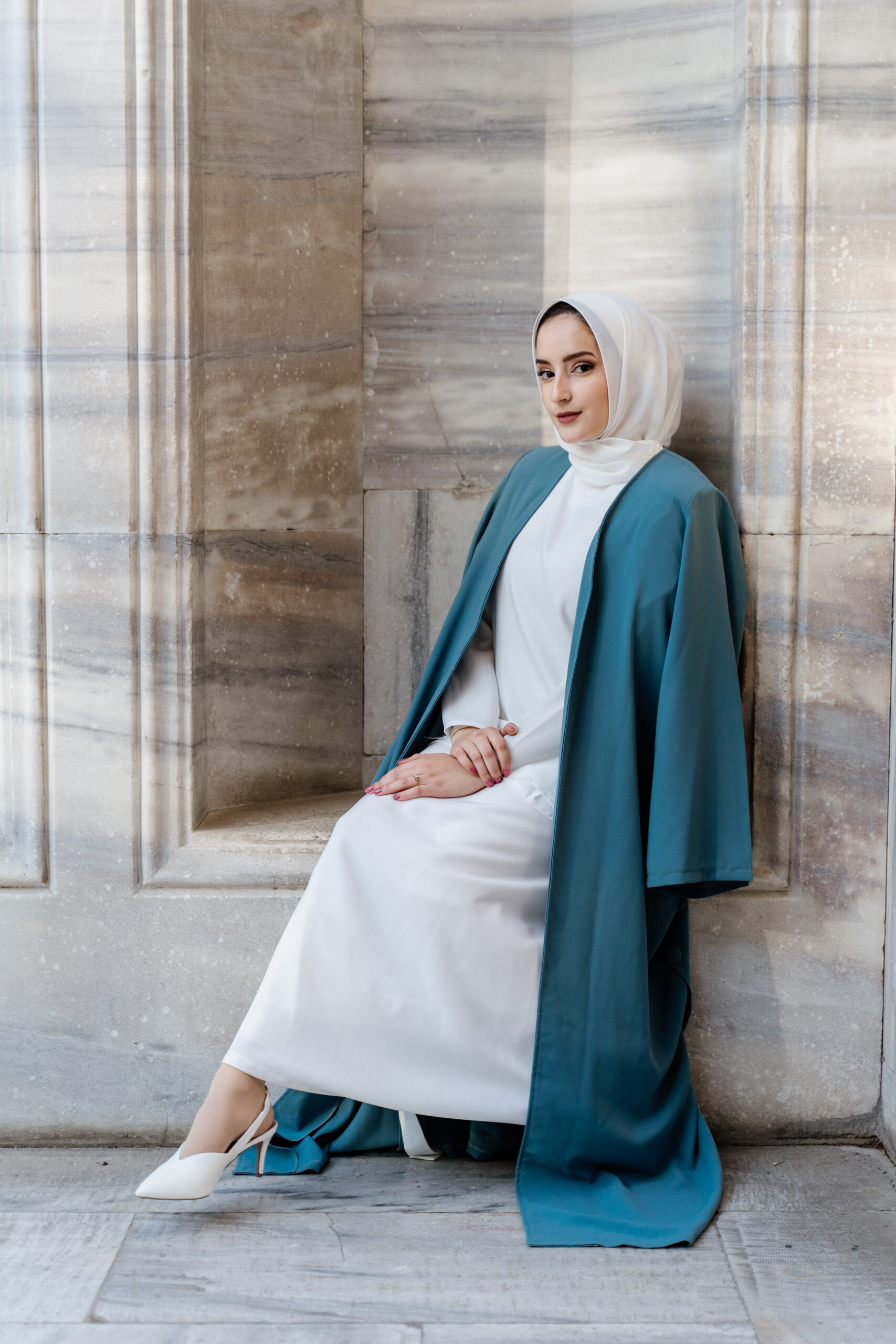 Woman in a white dress and teal coat seated beside marble columns
