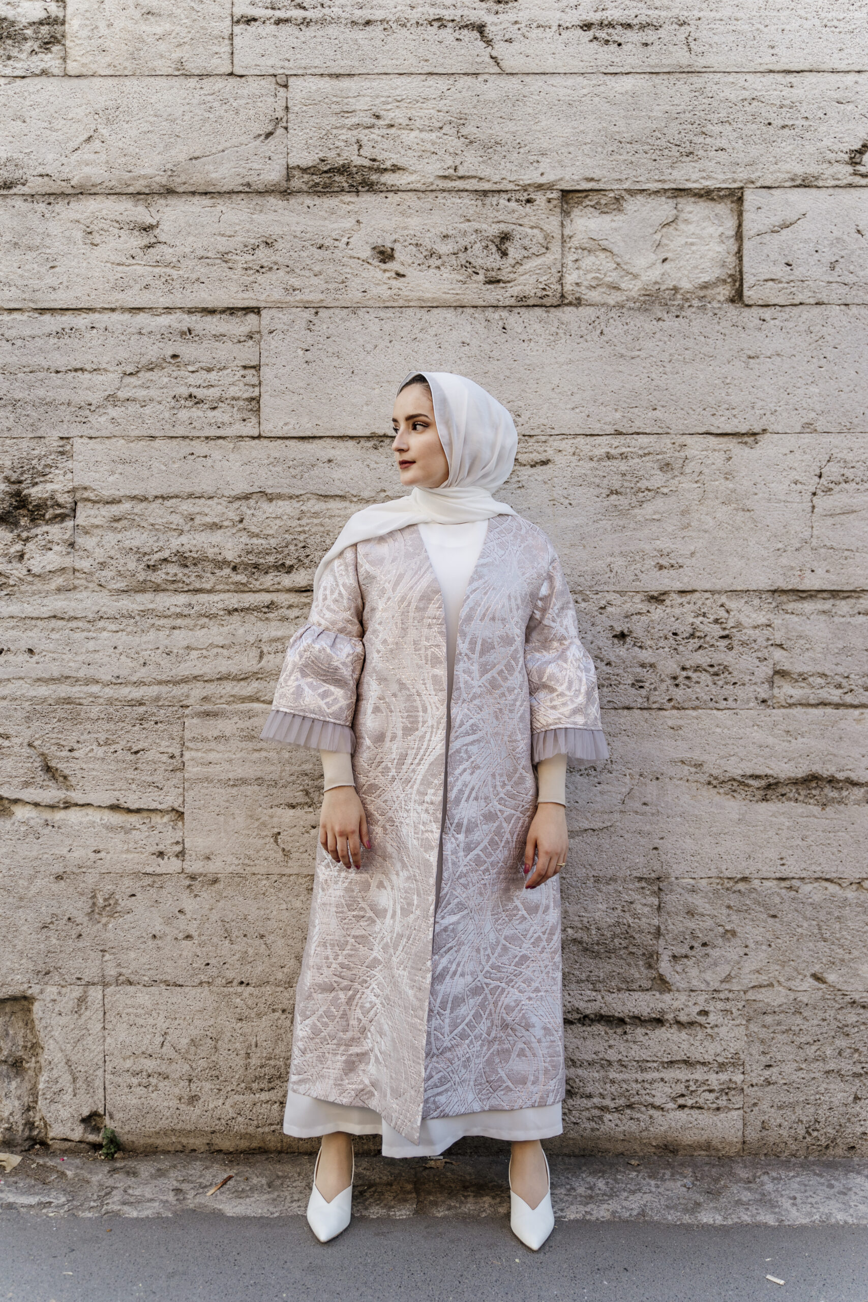 Woman in a pale patterned coat standing against a stone wall