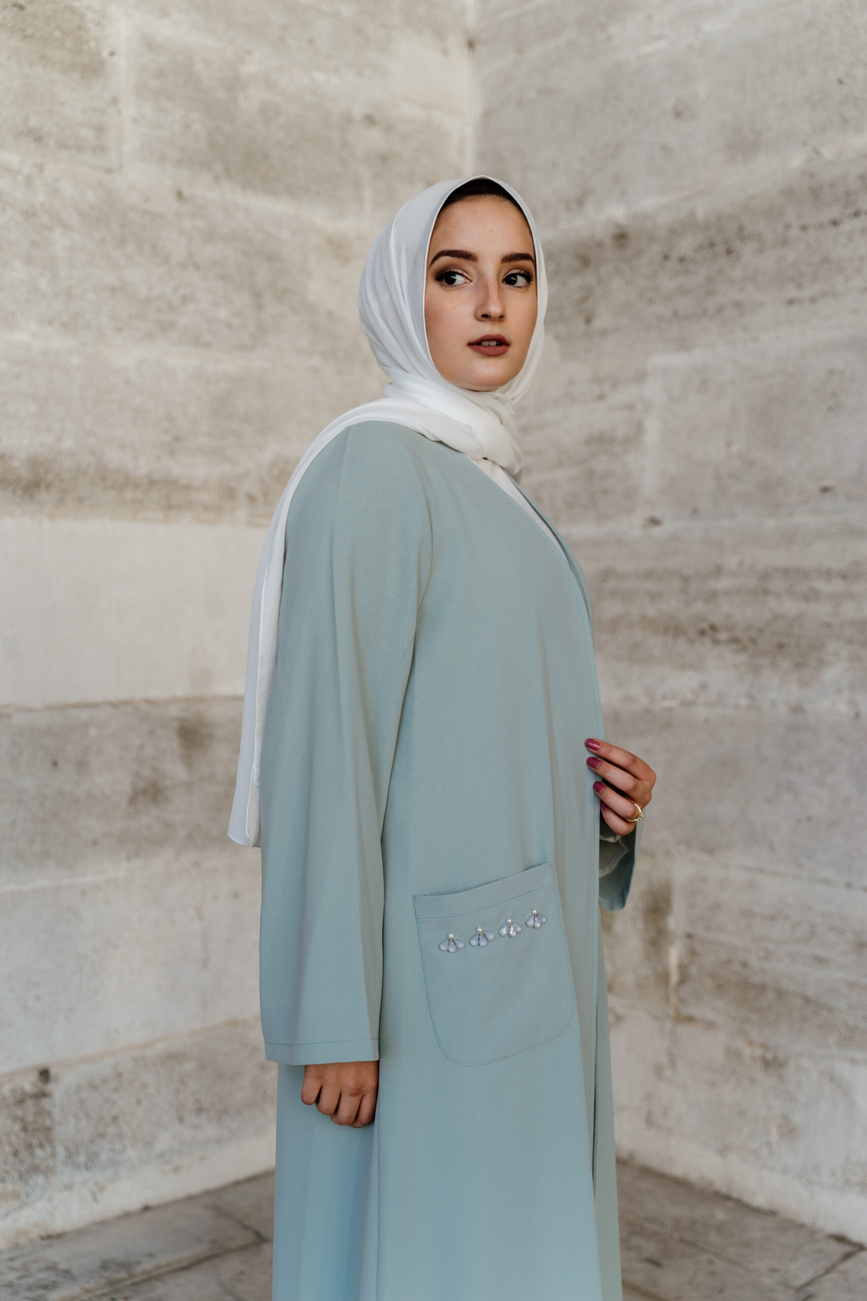 Woman in a light blue abaya standing by a stone wall